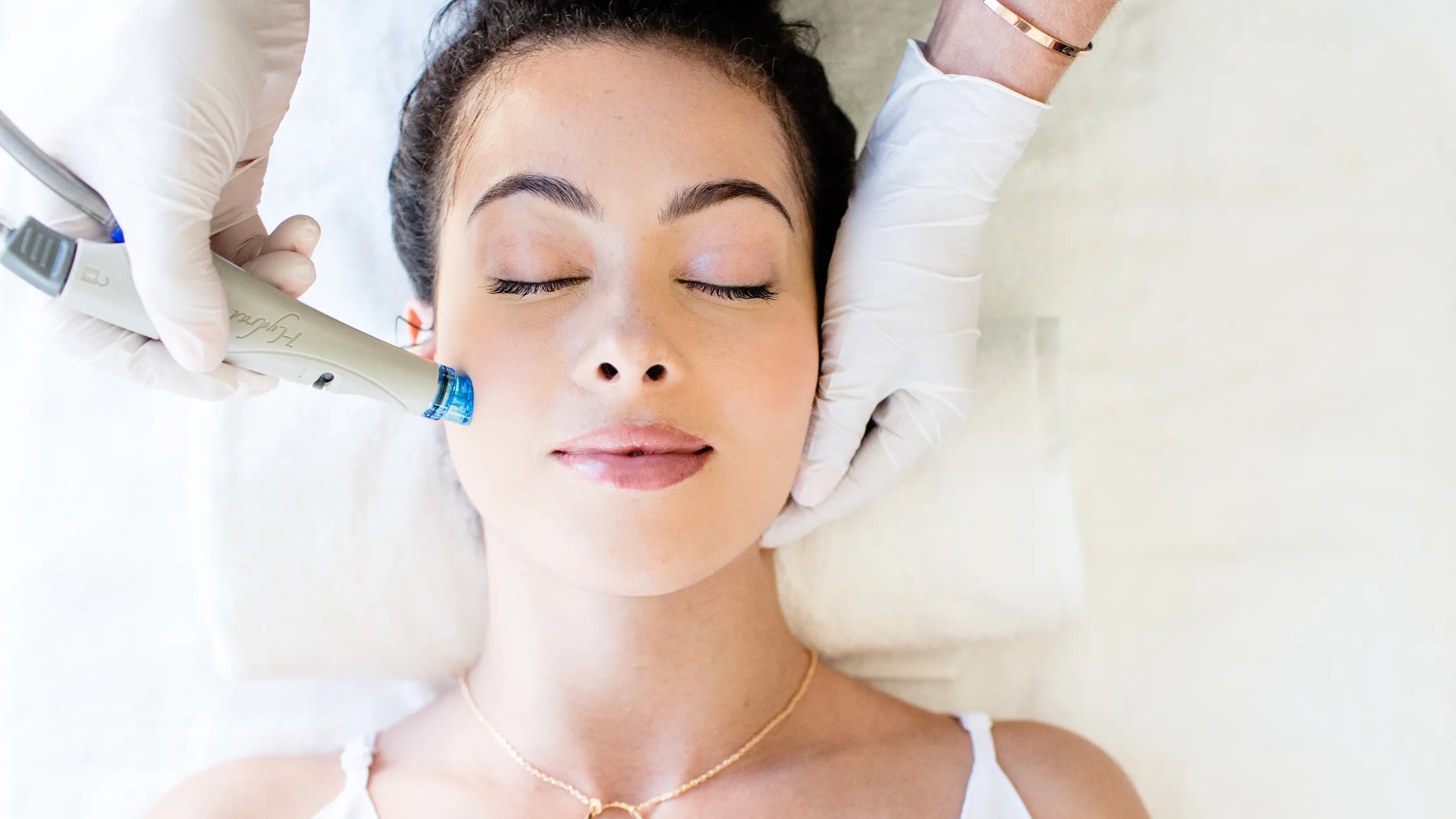 A woman with closed eyes lies on her back on a white towel, receiving a HydraFacial treatment. The aesthetician's gloved hands are visible, holding a HydraFacial wand to the woman's forehead. The woman appears relaxed, with her hair pulled back, during this skincare procedure.